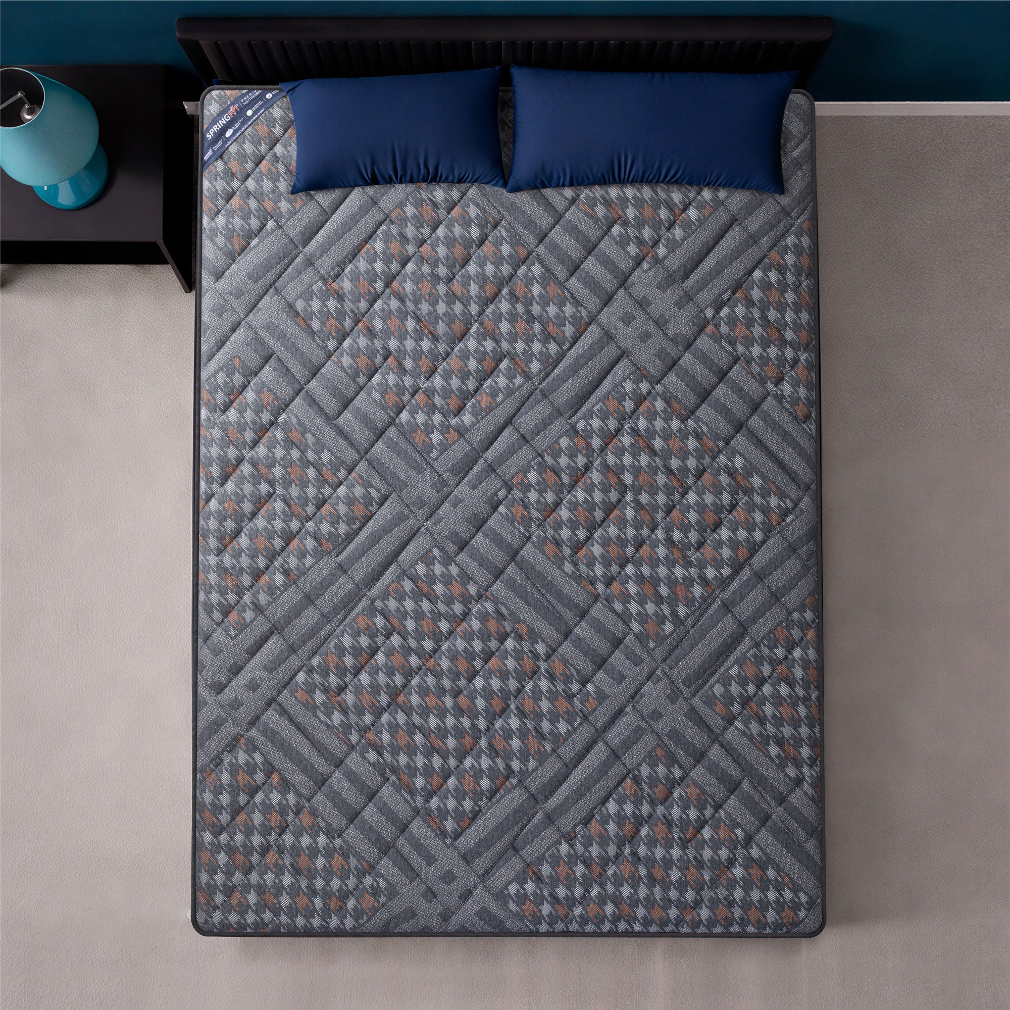 Overhead top-down view of the Springfit Betterspine Support mattress showing its full grey and brown houndstooth-patterned quilted fabric surface on a black bed with two navy blue pillows and a teal lamp on the nightstand