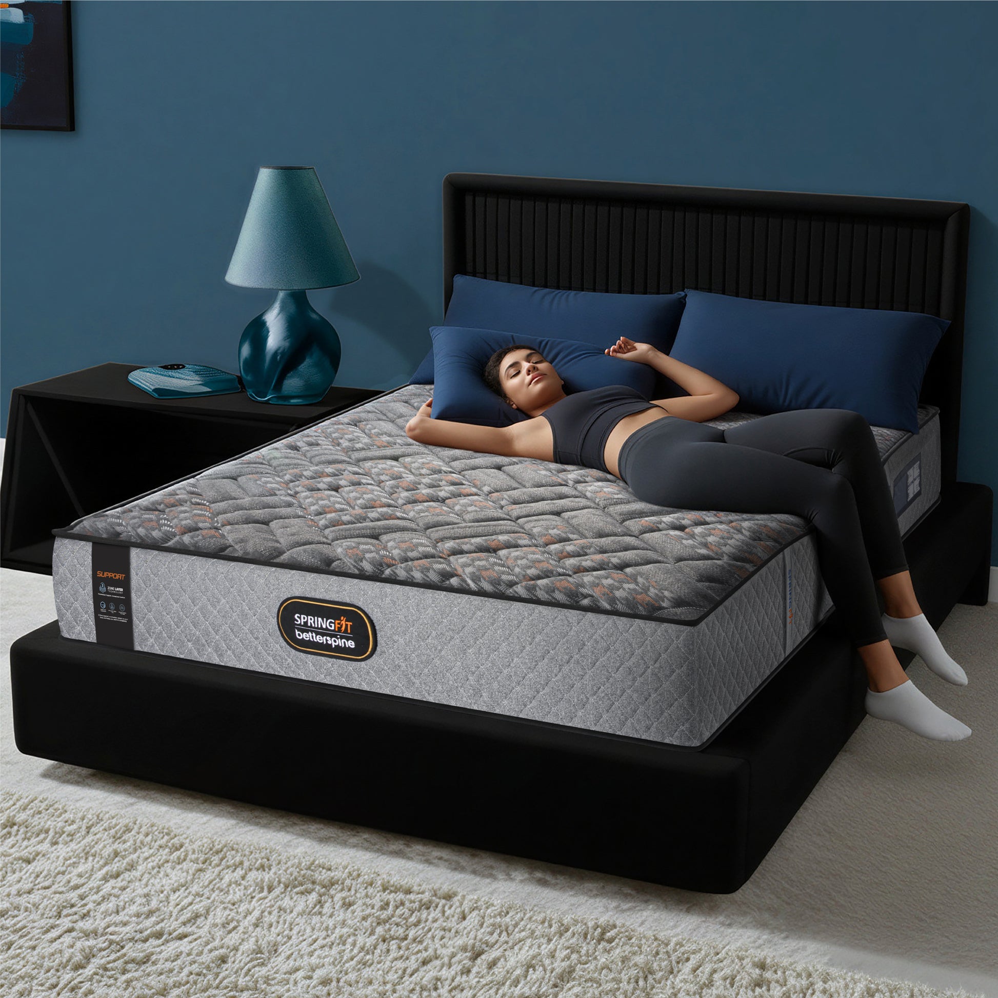 Springfit Betterspine Support mattress on a black platform bed with a tall dark channelled headboard in a navy blue bedroom, with a woman in grey activewear lying on her back in a relaxed sleeping position; teal table lamp and blue pillows visible