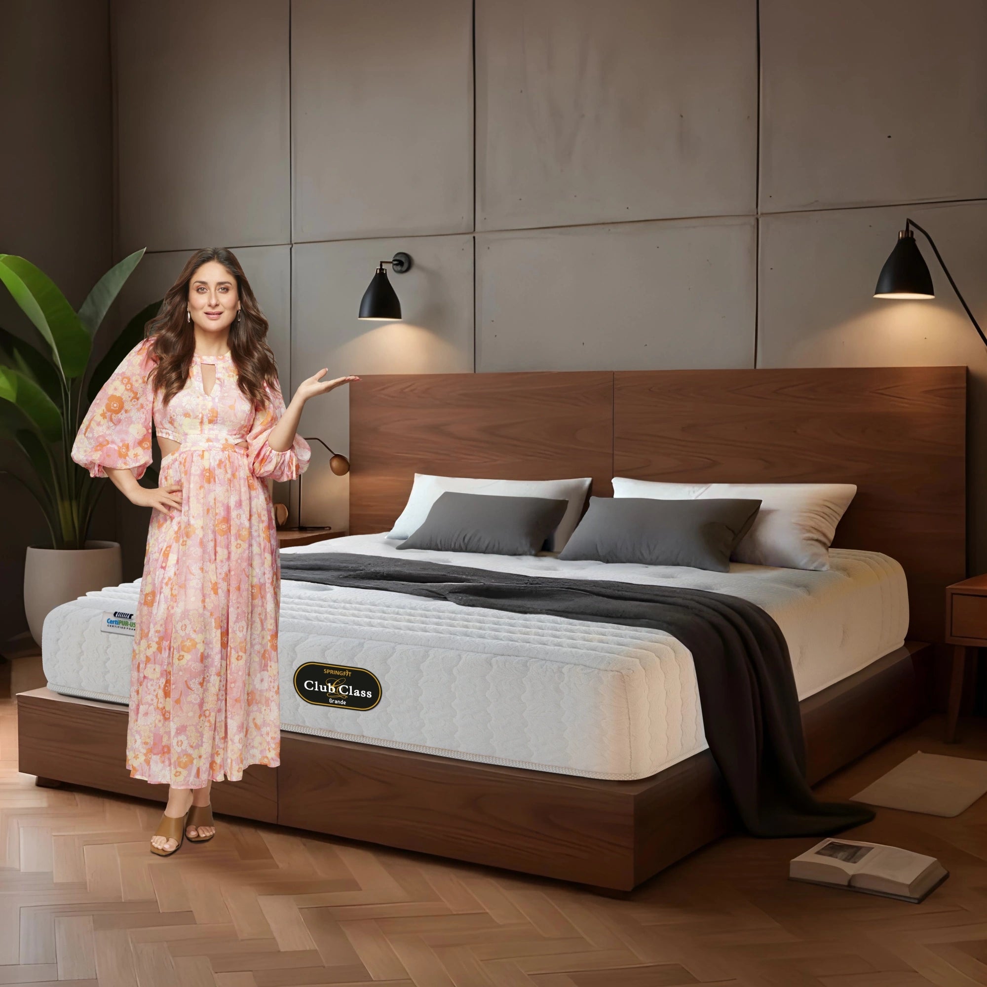 Springfit Club Class Grande luxury mattress lifestyle image with brand ambassador in pink floral dress gesturing toward mattress on walnut platform bed with grey pillows and dark throw in industrial-style bedroom with black wall sconces and indoor plant