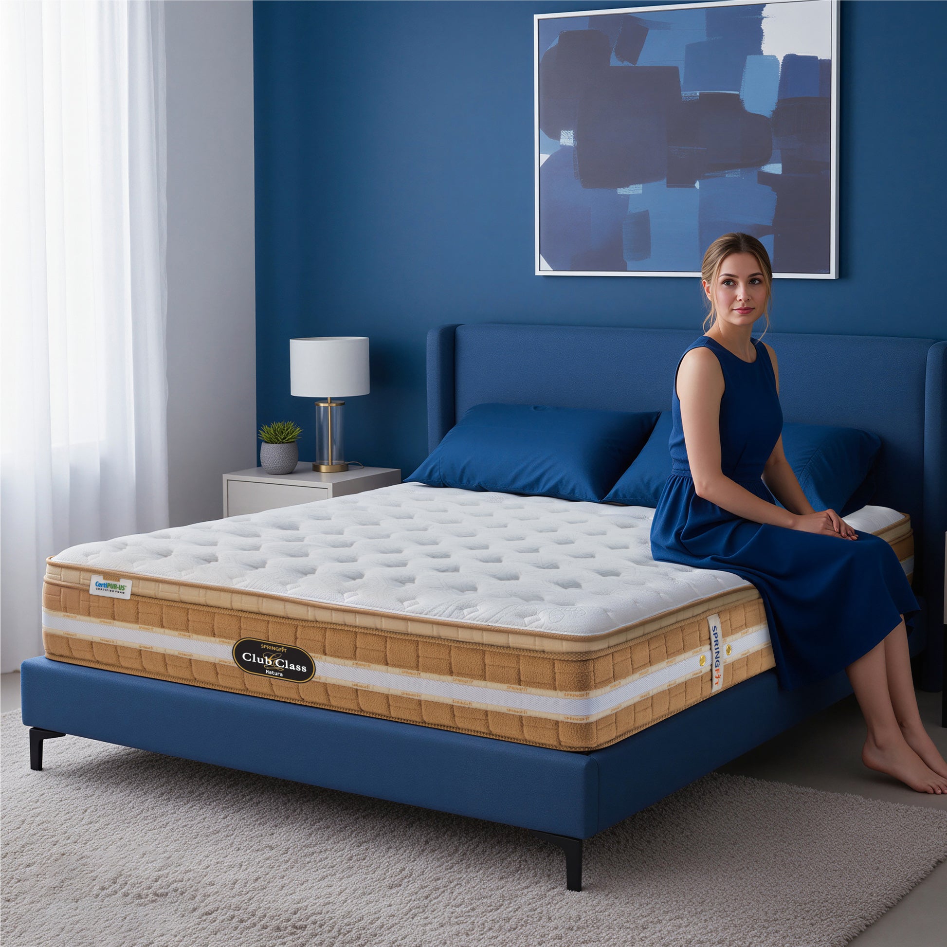 Springfit Club Class Natura luxury mattress lifestyle image with woman in navy blue dress seated on mattress edge on blue upholstered bed in navy blue accent bedroom with white lamp and abstract wall art