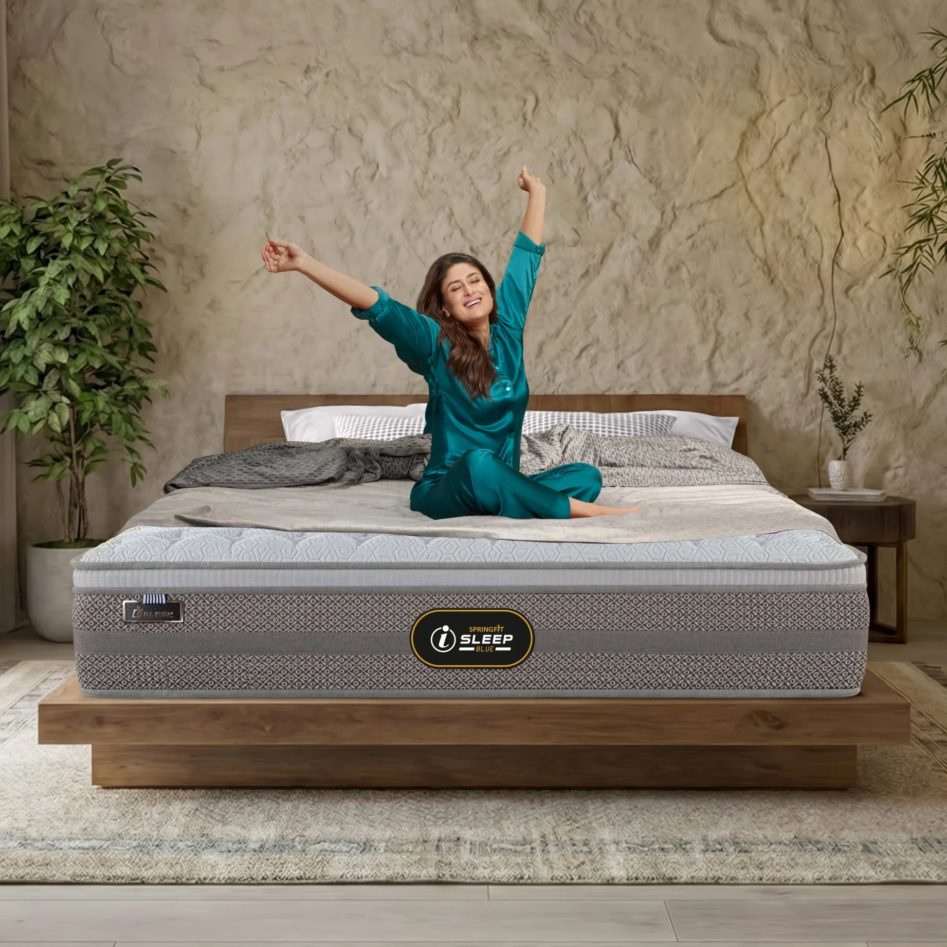 Springfit iSleep Blue mattress on a low wooden platform bed in a warm boho bedroom with textured plaster walls and indoor plants, with a brand ambassador in teal loungewear joyfully stretching her arms overhead