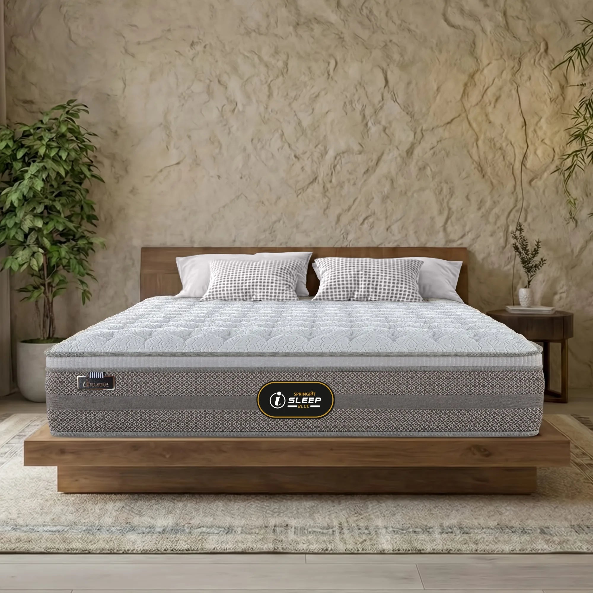 Front-facing view of the Springfit iSleep Blue mattress with a white diamond-quilted top and grey patterned fabric border, placed on a low natural wood platform bed in a warm boho bedroom with textured plaster walls and indoor plants
