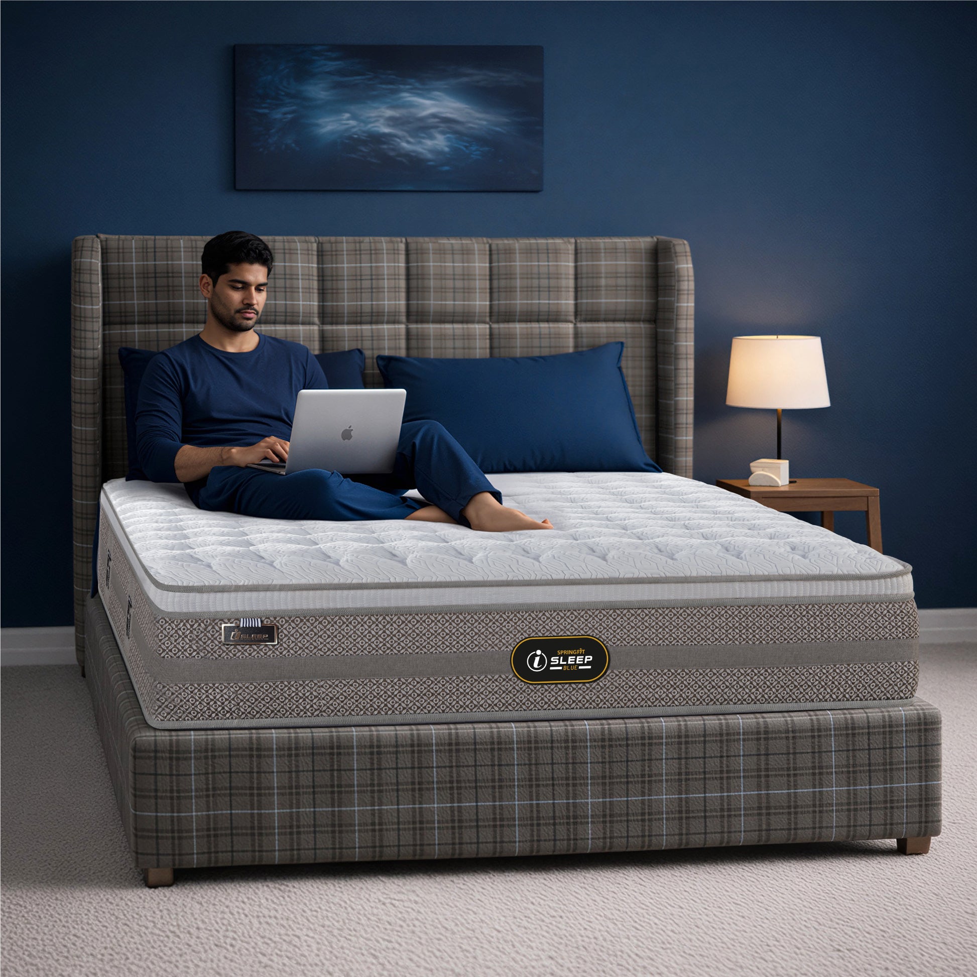 Springfit iSleep Blue mattress on a grey plaid upholstered wingback bed in a navy blue bedroom, with a man in blue loungewear working on a laptop while seated on the mattress