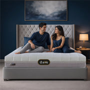 Springfit iSleep Krono mattress on a grey upholstered platform bed with a tall tufted wingback headboard in a navy blue bedroom, with a smiling couple in casual loungewear seated together on the mattress