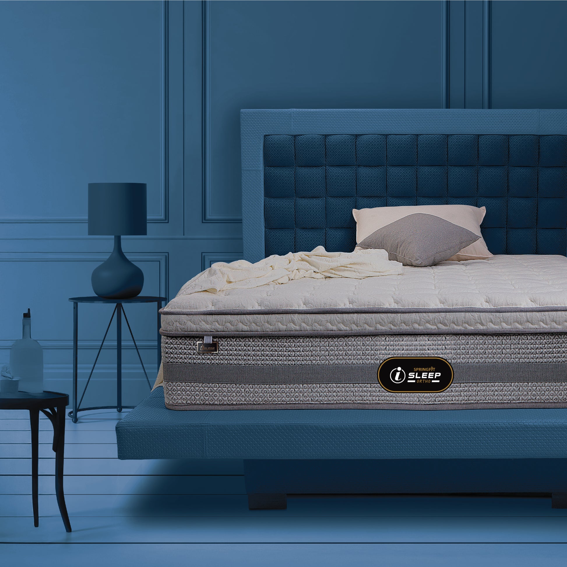 Springfit iSleep Ortho mattress displayed on a blue upholstered bed frame in a moody, dark-blue luxury bedroom with a side table lamp and decorative accents