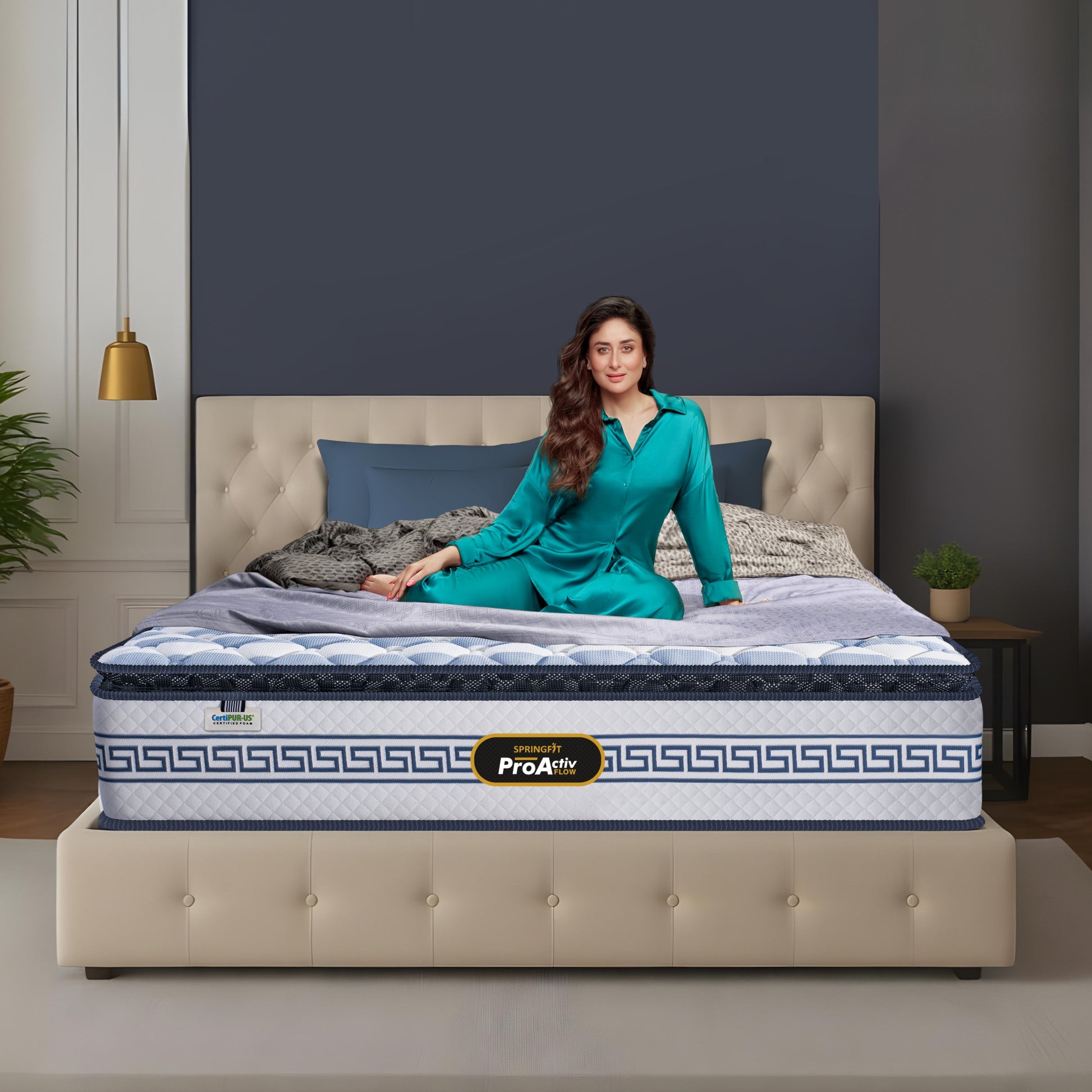 Springfit ProActiv Flow mattress lifestyle image with brand ambassador in teal outfit seated on bed with beige tufted headboard in blue-grey accent bedroom with gold lighting