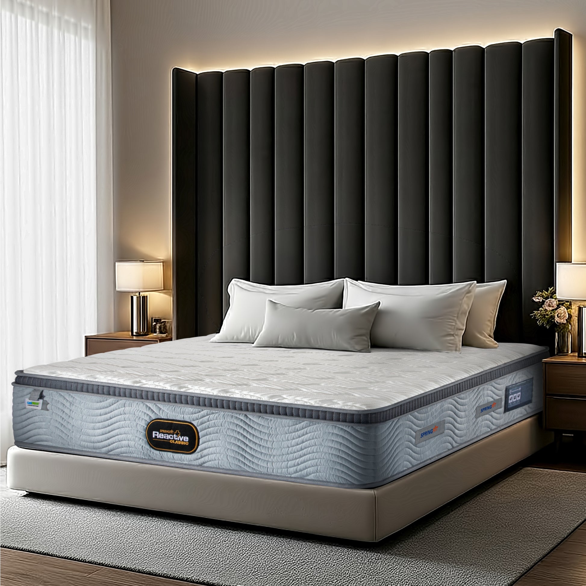 Angled side view of the Springfit Reactive Classic mattress with a white quilted pillow top and grey wave-patterned fabric border, on a beige upholstered platform bed in a luxury bedroom with a dramatic dark grey channelled headboard, sheer curtains, and table lamps