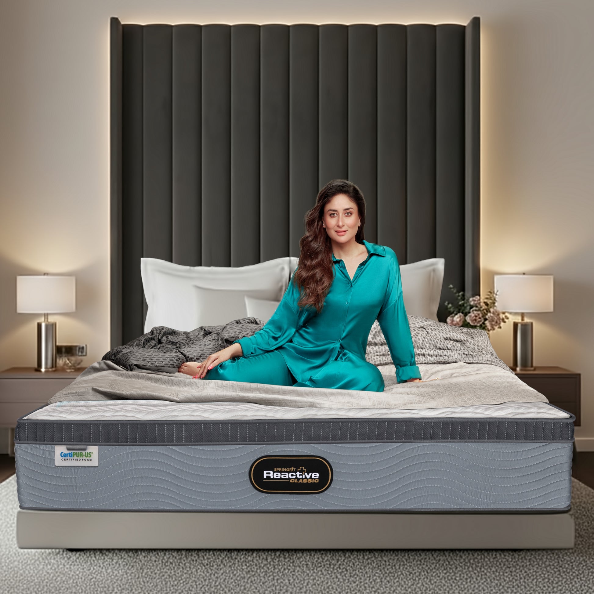 Springfit Reactive Classic mattress on a platform bed with a tall dark grey channelled velvet headboard in an elegant bedroom with silver cylinder lamps, with a brand ambassador in teal silk loungewear seated on the mattress; CertiPUR-US label visible on the side
