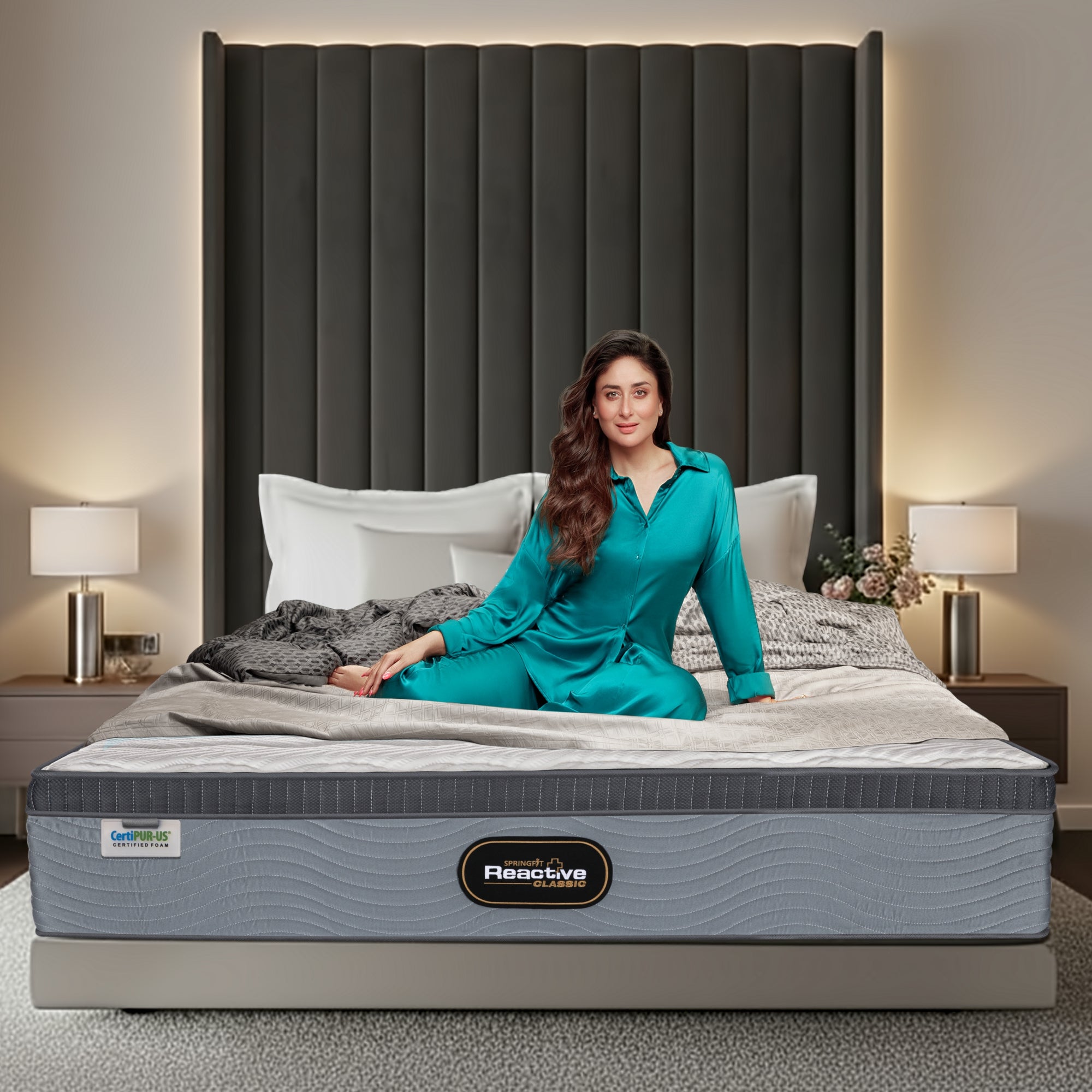 Springfit Reactive Classic mattress on a platform bed with a tall dark grey channelled velvet headboard in an elegant bedroom with silver cylinder lamps, with a brand ambassador in teal silk loungewear seated on the mattress; CertiPUR-US label visible on the side