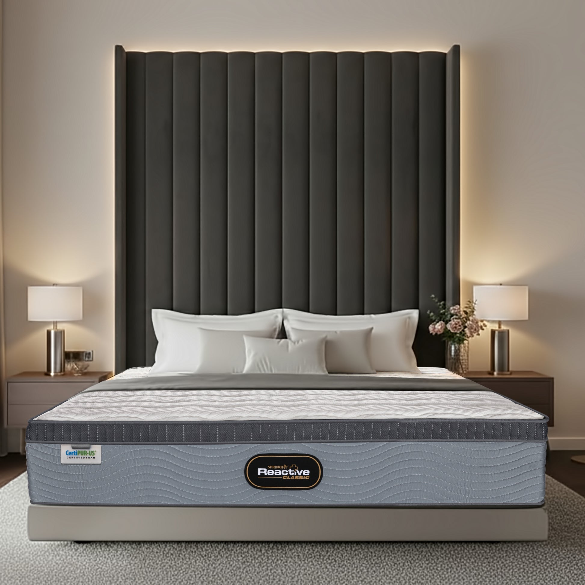 Front-facing view of the Springfit Reactive Classic mattress with a white wave-quilted top, dark grey border trim, and grey wave-textured fabric base, on a beige platform bed flanked by silver cylinder table lamps and a tall dark channelled headboard