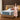 Springfit Reactive Dual Memory mattress lifestyle image with brand ambassador in pink floral dress gesturing toward mattress on beige platform bed in warm wood-paneled bedroom with twin side lamps