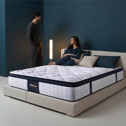 Springfit Reactive Gold mattress lifestyle image with couple in casual outfits — woman seated and man standing — on beige upholstered bed with teal and beige pillows in dramatic dark teal accent bedroom with floor lamp