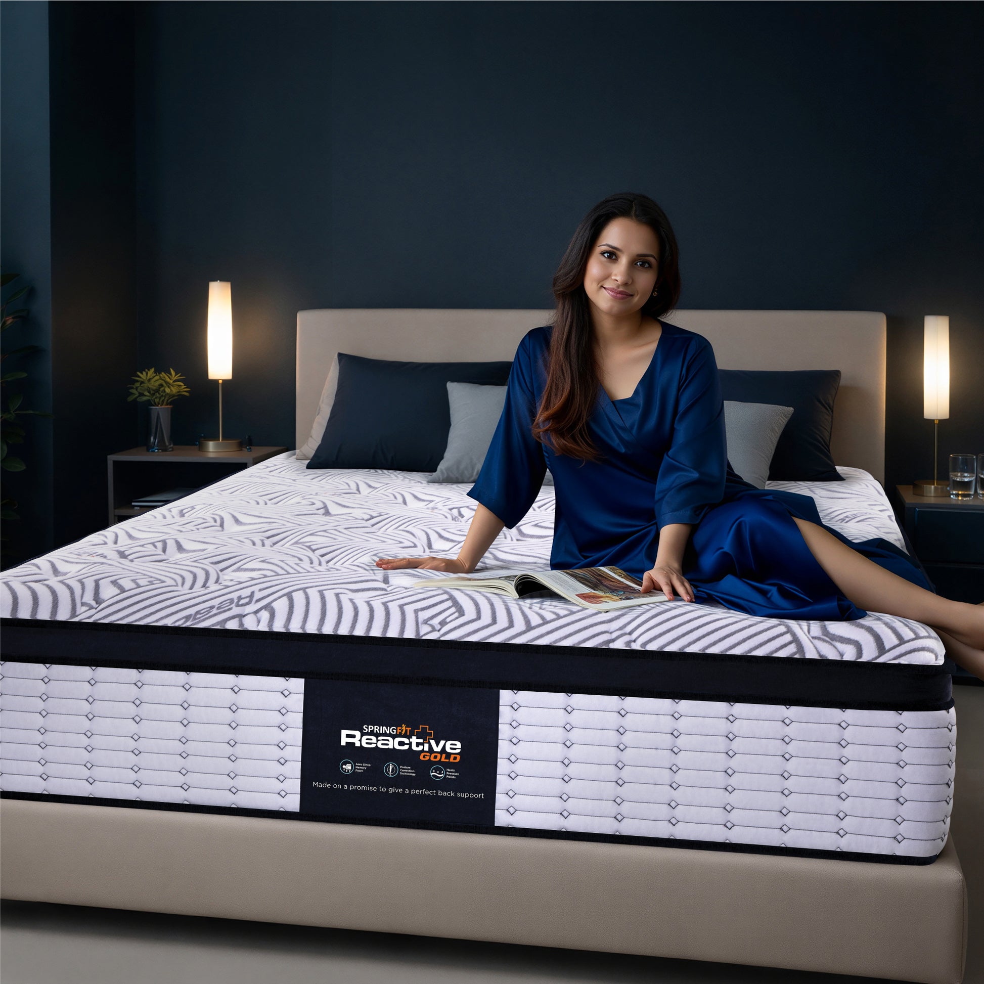 Springfit Reactive Gold mattress lifestyle image with woman in navy blue satin outfit seated on mattress with open magazine on beige platform bed in dark teal bedroom with twin floor lamps and indoor plant