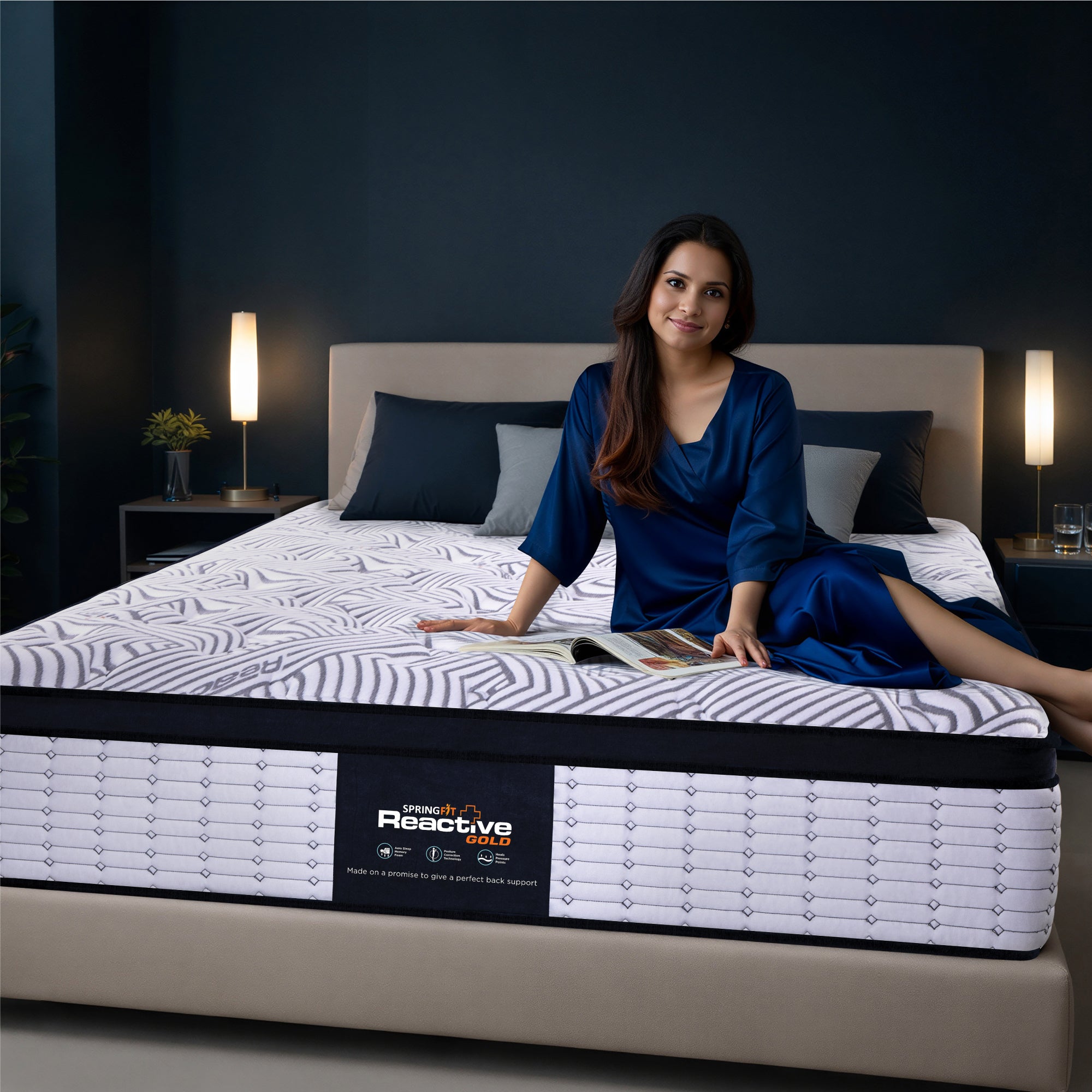 Springfit Reactive Gold mattress lifestyle image with woman in navy blue satin outfit seated on mattress with open magazine on beige platform bed in dark teal bedroom with twin floor lamps and indoor plant