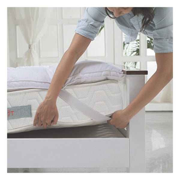 Woman securing the elastic corner strap of a Springfit mattress topper onto a mattress placed on a white wooden bed frame in a bright bedroom